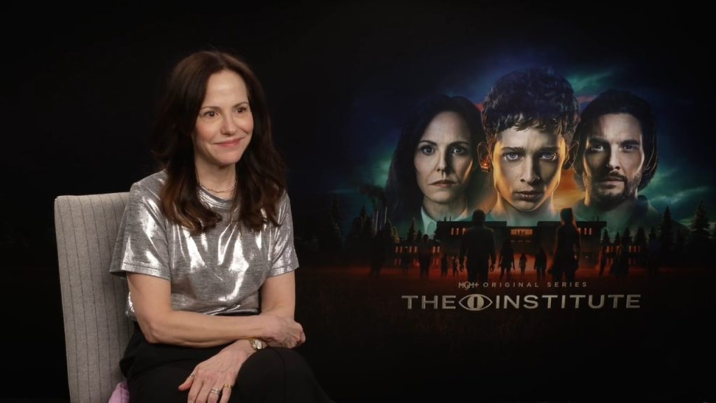 A woman sits on a chair next to a promotional poster for the MGM original series "The Institute," featuring three characters and the show's title.