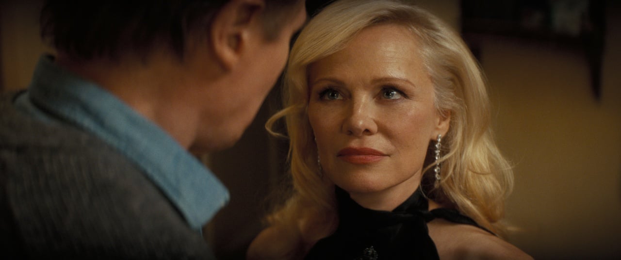 A blonde woman with wavy hair and dangling earrings looks intently at a man in a dimly lit indoor setting.
