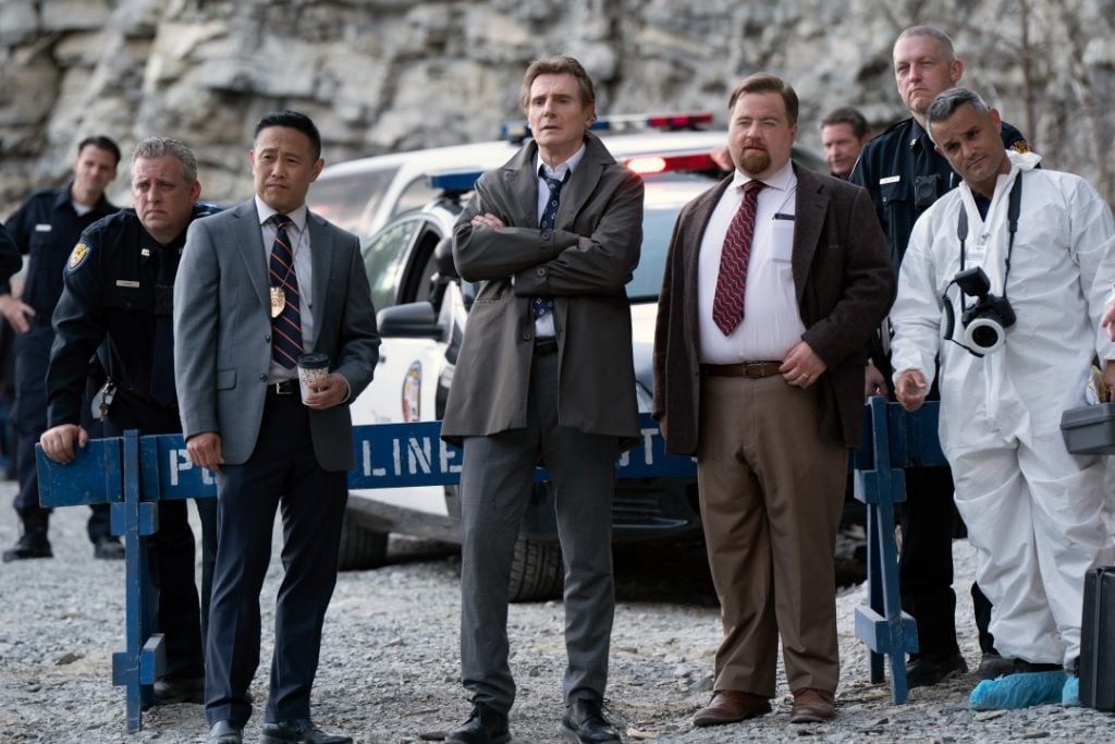 A group of detectives, police officers, and a forensic investigator stand in front of a police car at a cordoned-off outdoor crime scene.