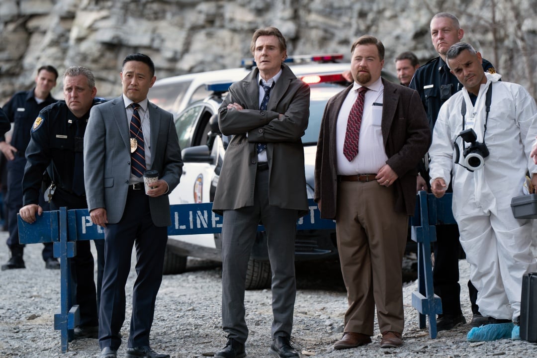 A group of detectives, police officers, and a forensic investigator stand in front of a police car at a cordoned-off outdoor crime scene.