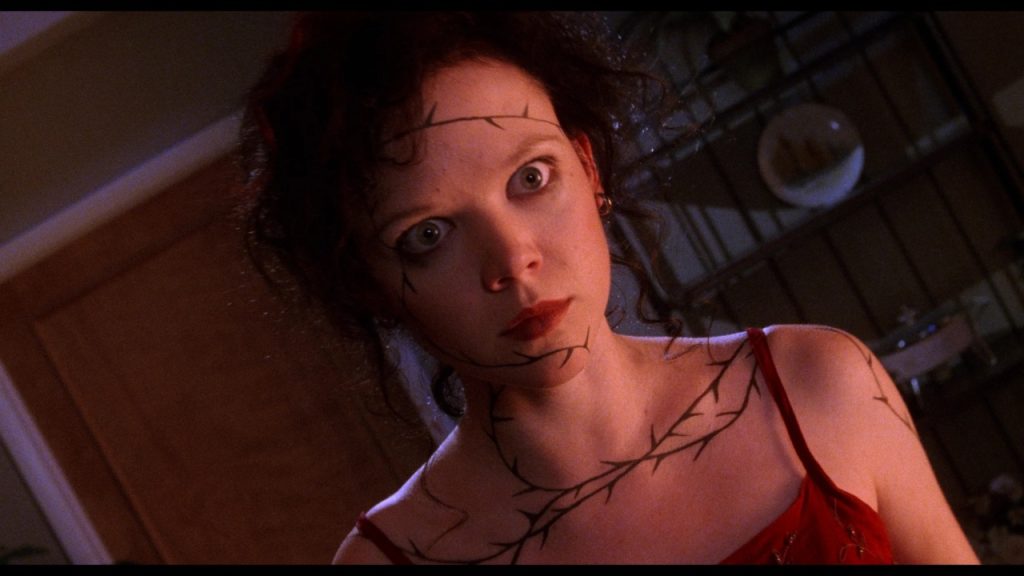 A woman with tangled hair, wide eyes, and black vine-like markings on her skin stands indoors, illuminated by reddish light.