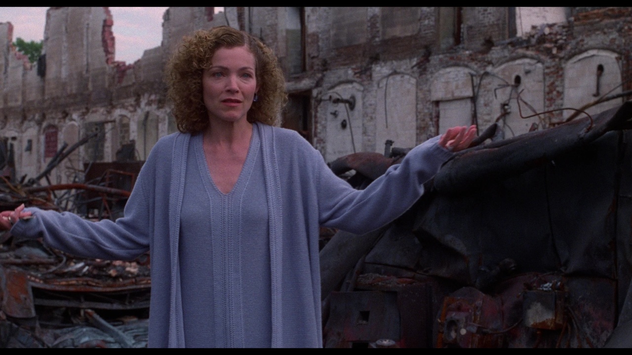 A woman with curly hair stands in front of a backdrop of destroyed buildings and debris, wearing a light blue sweater with her arms slightly raised.