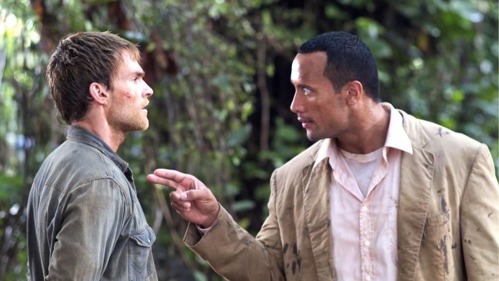 Two men stand outdoors facing each other; the man on the right gestures with his hand while speaking sternly to the man on the left. Lush greenery is visible in the background.