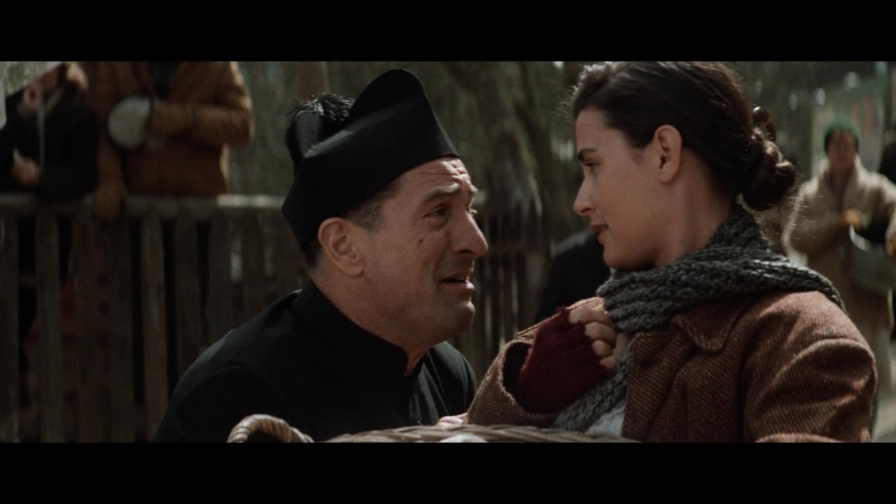 A man in a black hat and dark clothing looks up at a woman in a brown coat and scarf; they appear to be speaking closely outdoors.
