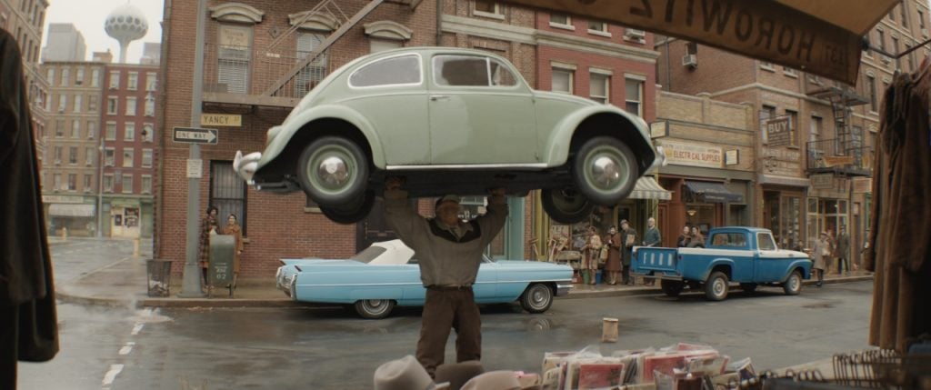 A man lifts a green Volkswagen Beetle above his head on a city street while bystanders watch; vintage cars and storefronts are visible in the background.