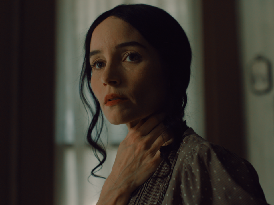 A woman with dark hair and a neutral expression holds her hand to her neck, standing indoors in soft, natural light.