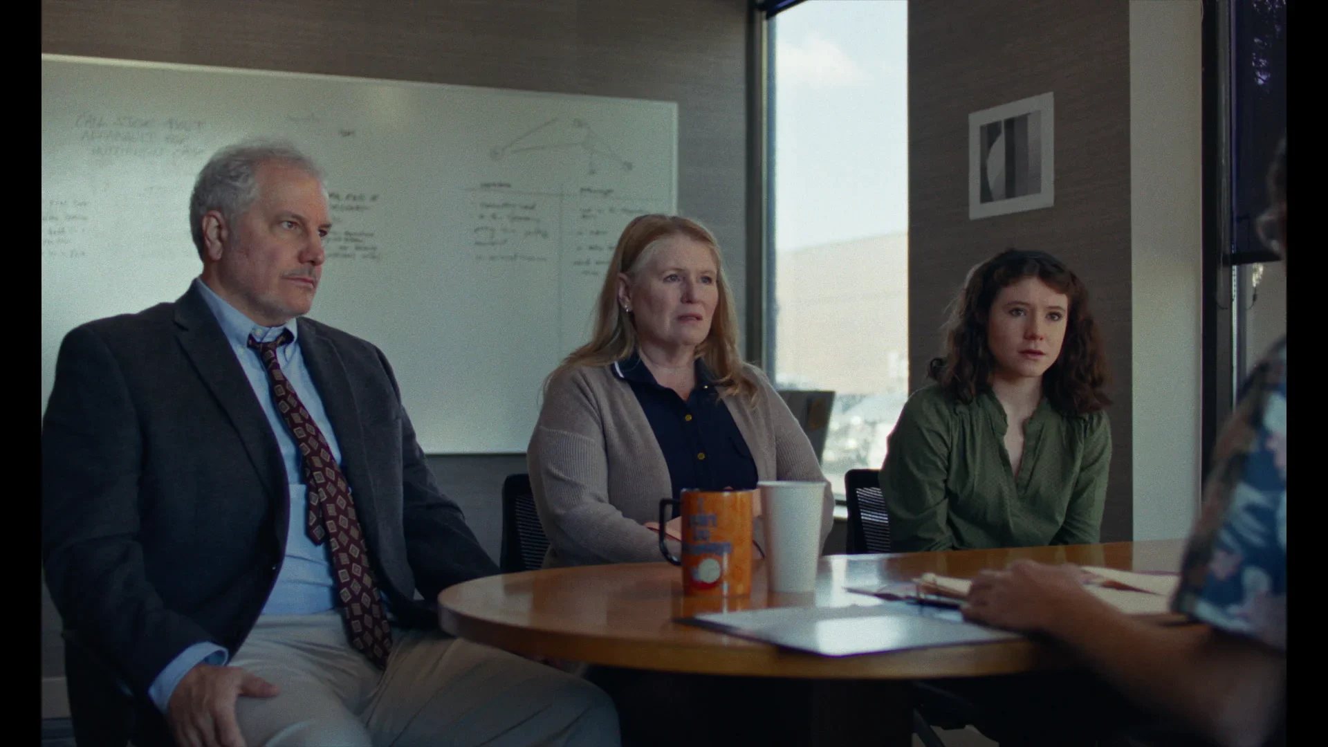 Three people sit at a round table in an office, facing another person whose back is visible; a whiteboard and window are in the background.
