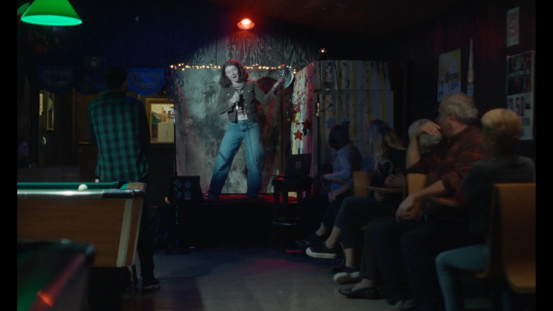 A performer with a guitar sings on a small stage under red lighting, while an audience sits in rows and a person stands near a pool table in the foreground.