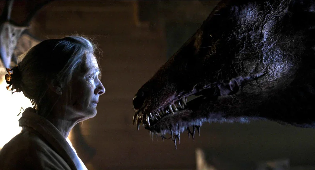 An older woman faces a large, menacing dinosaur-like creature in a dimly lit room.