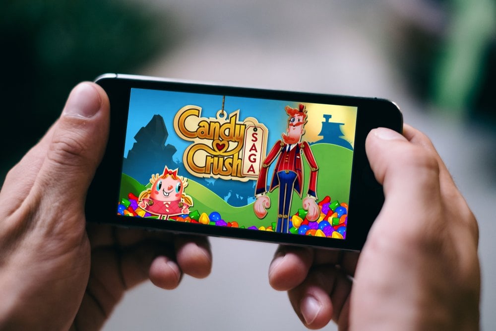 A person holds a smartphone displaying the Candy Crush Saga game start screen with two animated characters and colorful candy graphics.