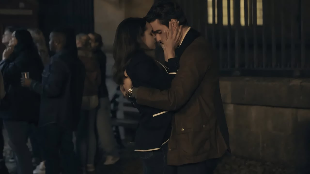 A man and a woman embrace and kiss outside at night, with other people gathered in the background.