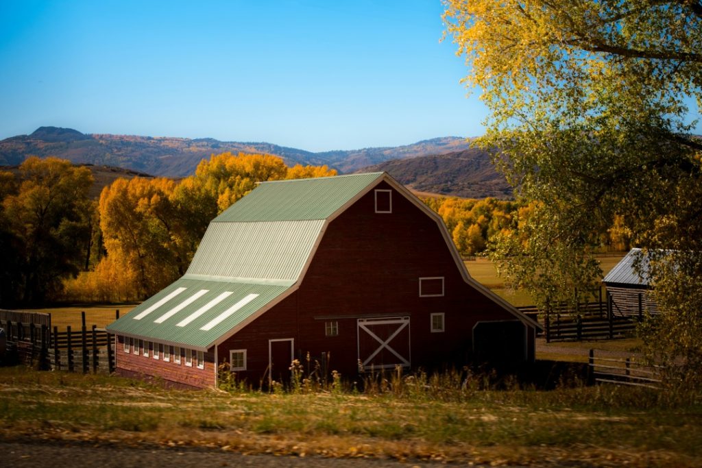 A red barn with a green roof sits in a rural landscape with autumn trees and hills in the background under a clear blue sky.
