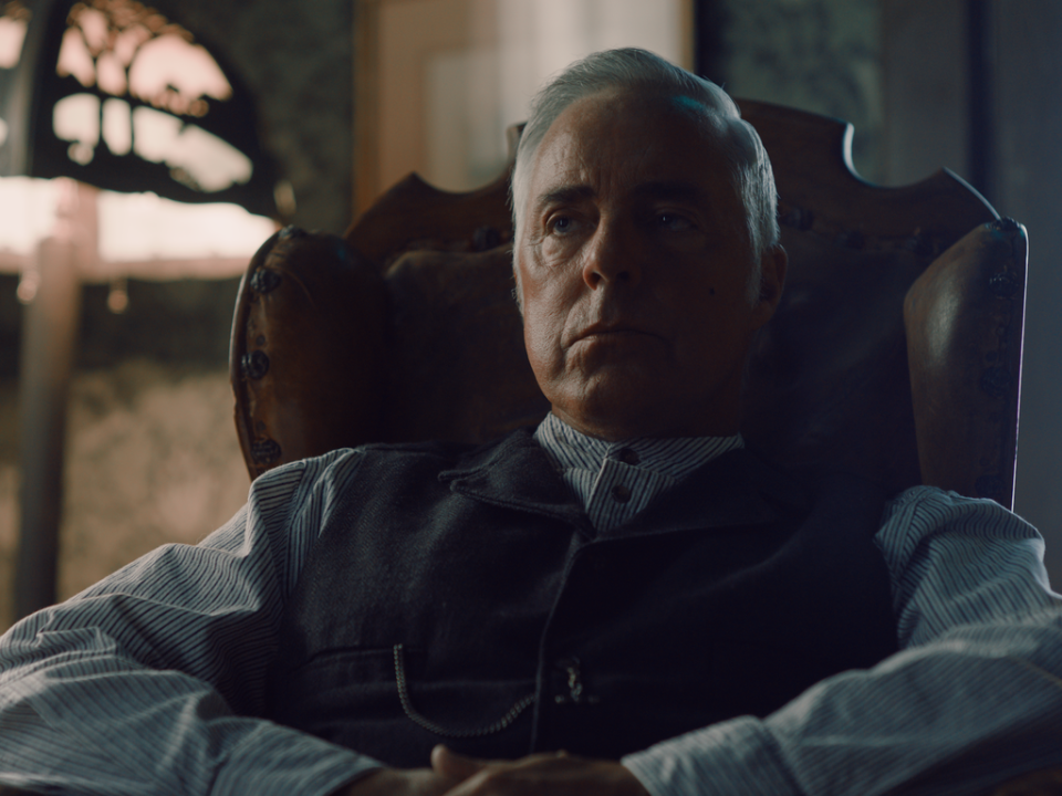An older man with gray hair sits in a leather chair, wearing a striped shirt and dark vest, in a dimly lit room.
