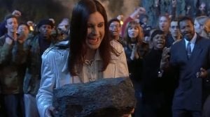 A person in a white jacket holds a large rock while a crowd of people stands behind, applauding and watching intently at night.