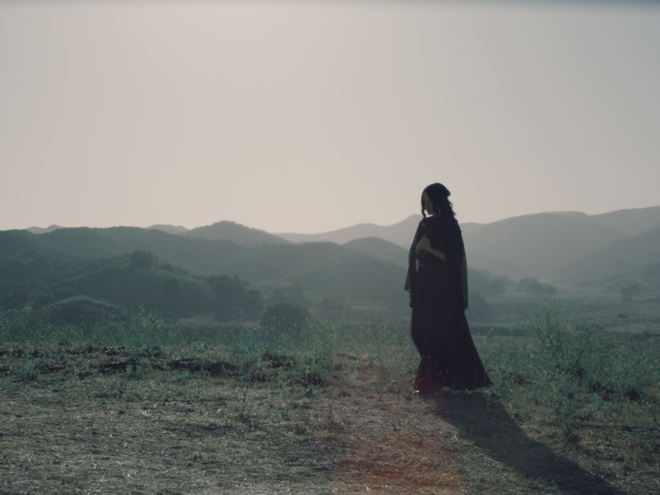 A person in dark clothing stands on a grassy field with hills in the background, under a bright, hazy sky.