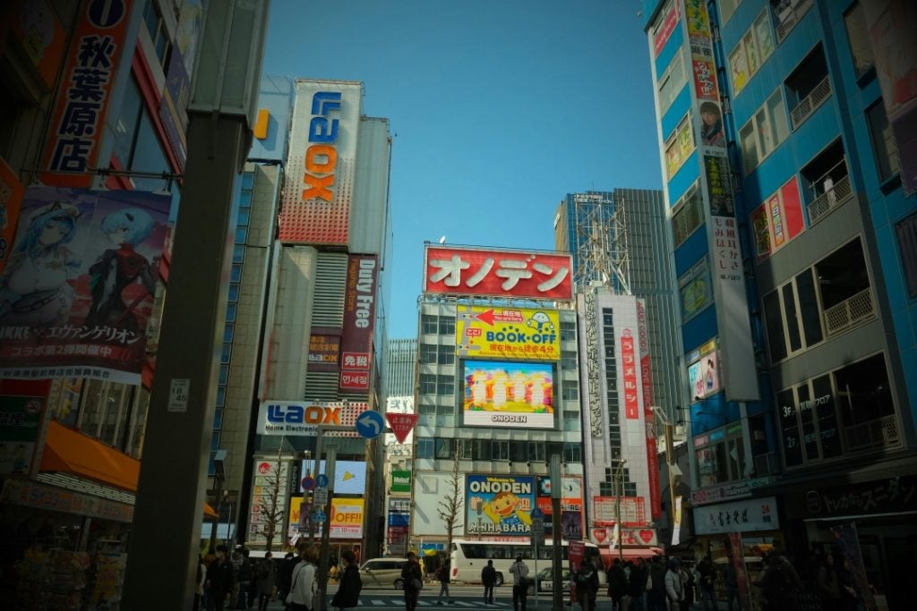 Street view of a busy urban area in Japan with tall buildings covered in colorful advertisements and signs in Japanese, and people walking below.