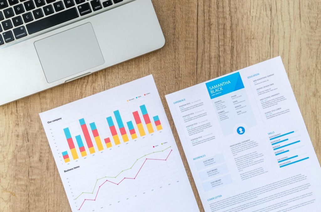 A laptop, a colorful bar and line graph printout, and a resume with blue highlights are arranged on a wooden desk.