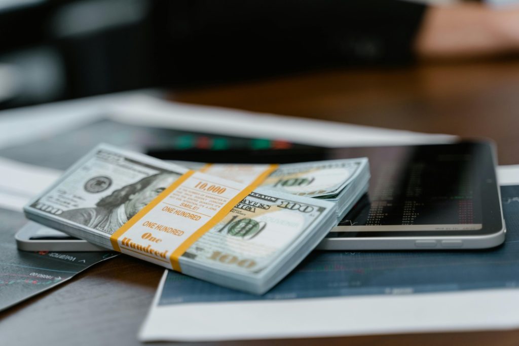 A stack of $100 bills in a paper band rests on a tablet displaying financial data, atop a desk with printed charts.