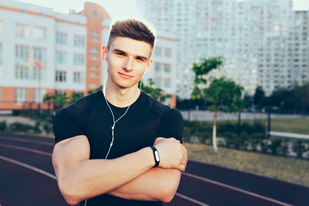 A young man in athletic wear stands on an outdoor running track with his arms crossed, wearing earphones and a fitness tracker, with buildings in the background.