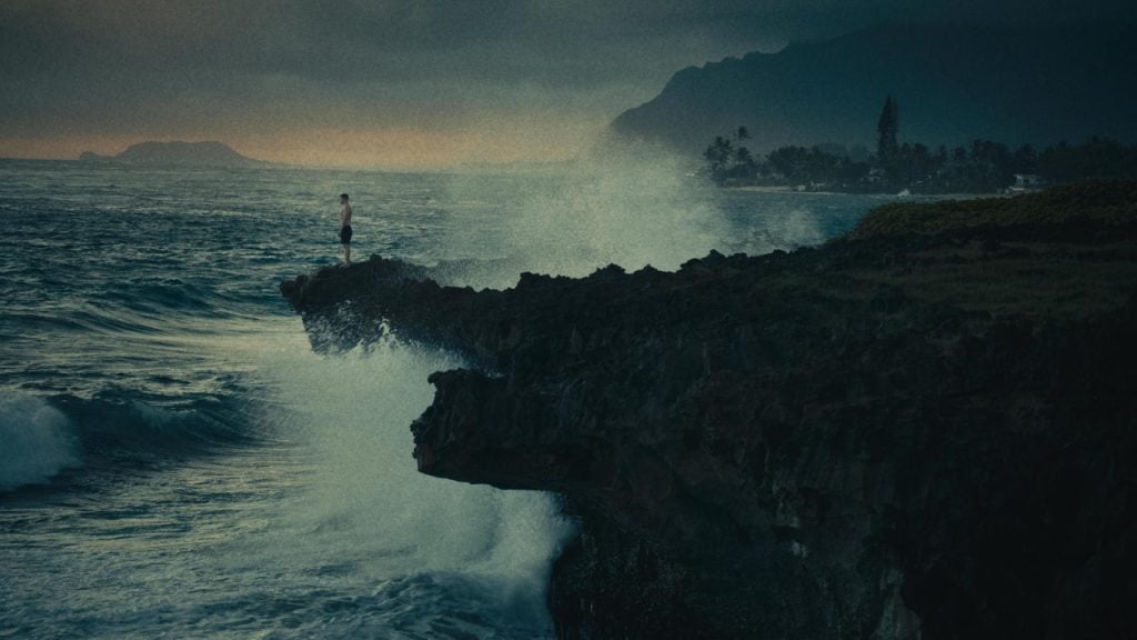 A person stands on a rocky cliff above crashing ocean waves, with mountains and a coastal town visible in the background under a cloudy sky.