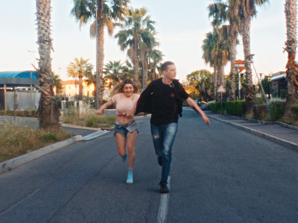 Two people, a woman and a man, run down a palm tree-lined street on a sunny day. The woman is barefoot and smiling, while the man leads her by the hand.