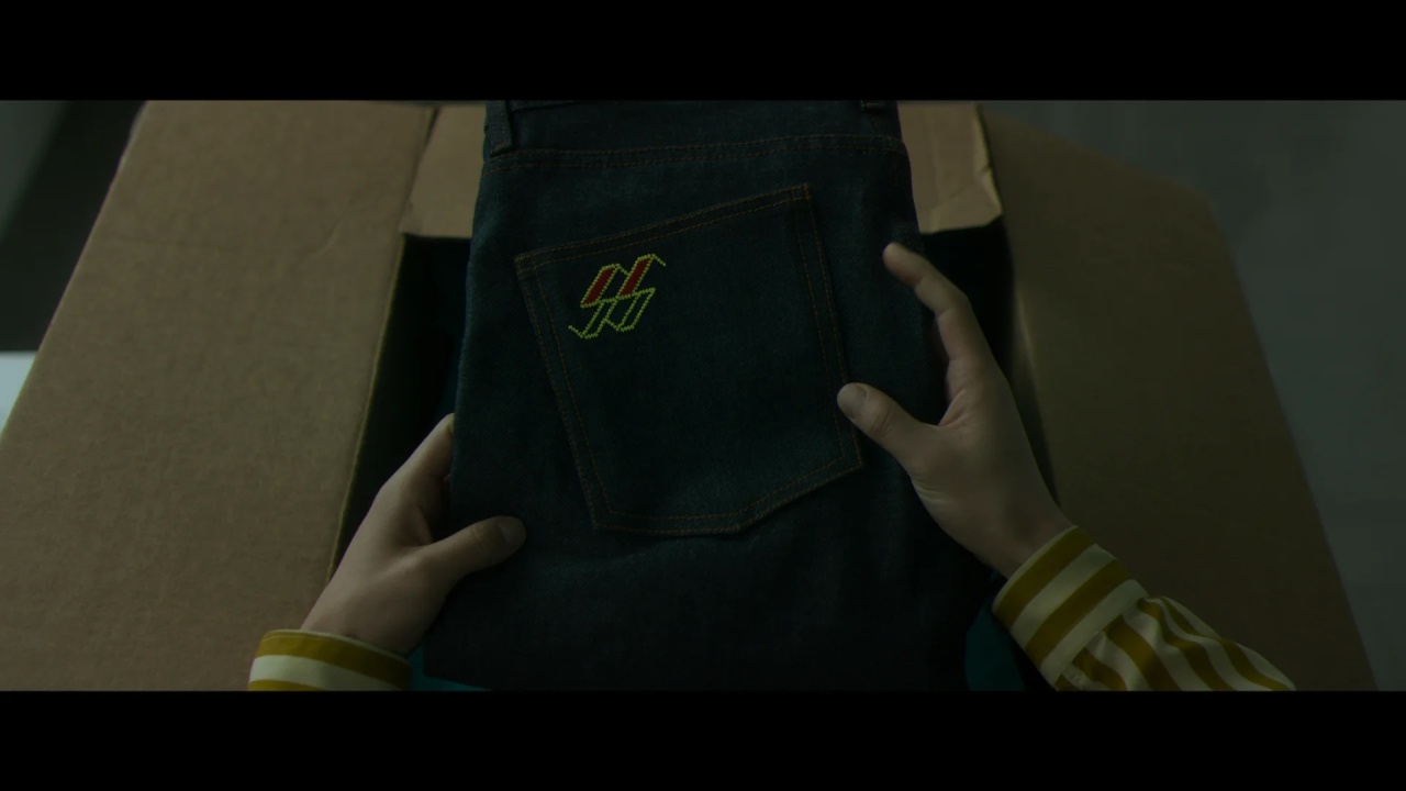 A person holds a pair of dark jeans with a colorful embroidered logo on the back pocket, viewed from above.