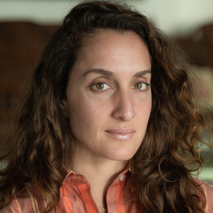 A woman with long, wavy brown hair and a neutral expression, wearing an orange striped shirt, looks directly at the camera.