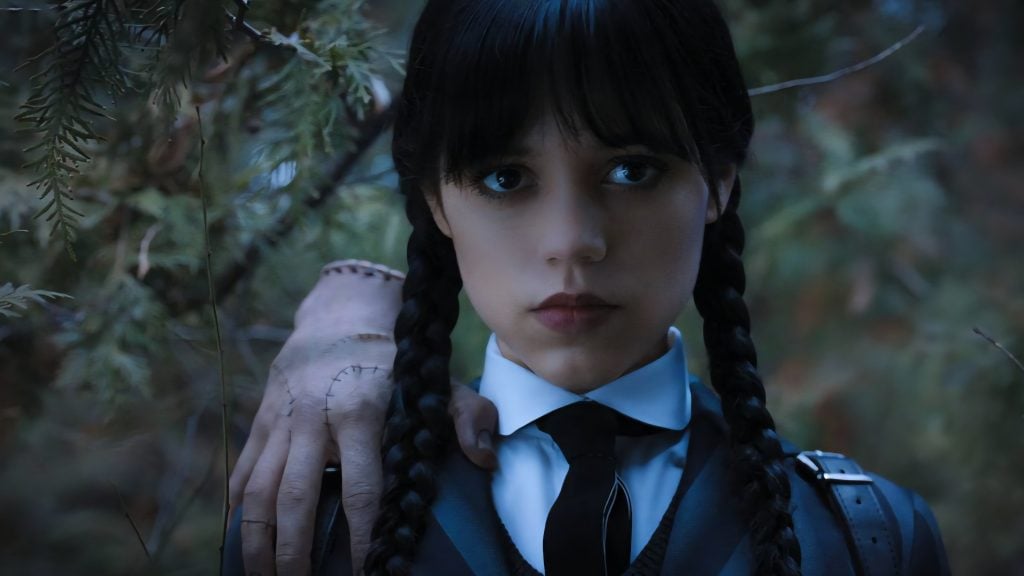 A girl with braided hair and a serious expression stands in a forest, wearing a school uniform. A disembodied hand rests on her shoulder.