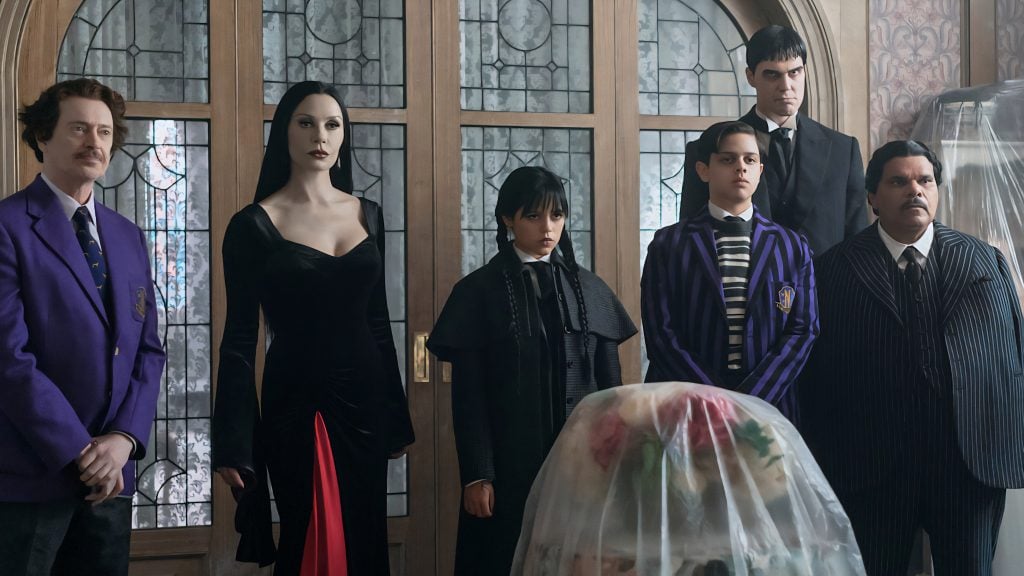 Six people stand indoors facing forward, dressed in dark or formal clothing, with a large covered object in front of them and ornate glass doors in the background.