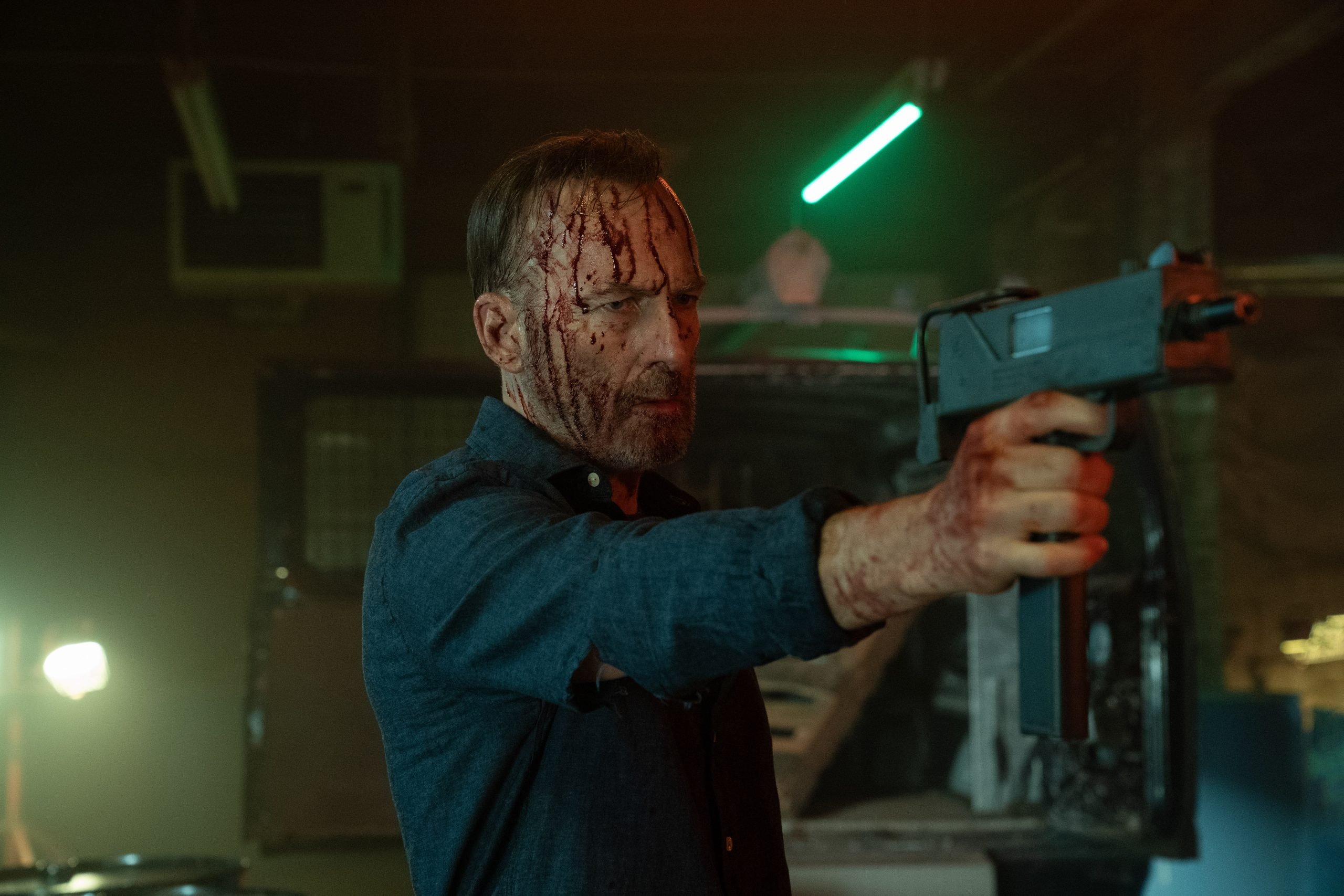 A man with a bloodied face and shirt points a submachine gun in a dimly lit, industrial setting.