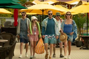 Five people in casual summer clothes walk outdoors, some wearing sunglasses and hats, with yellow umbrellas and patio furniture in the background.