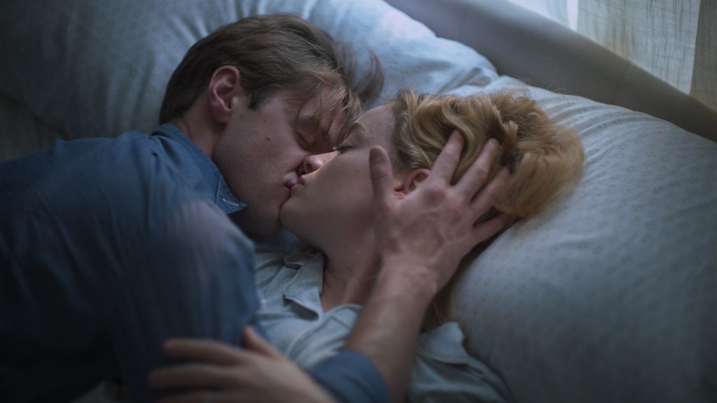 A man and woman lying in bed, kissing closely, with the man's hand gently holding the woman's head.