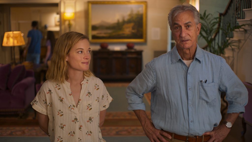 A young woman and an older man stand indoors, facing the camera. The woman smiles slightly while the man looks serious. The background shows a furnished living room and a painting on the wall.