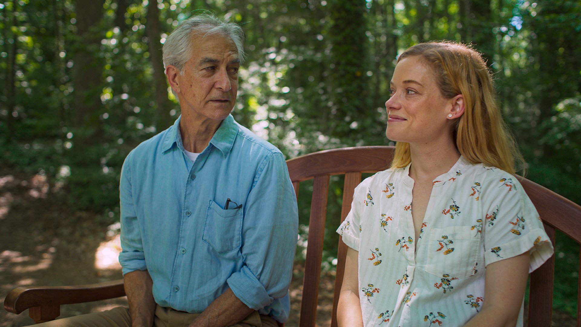 An older man and a younger woman sit on a wooden bench outdoors, facing each other, surrounded by trees and greenery.