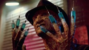 A burned man wearing a hat holds up a glove with syringes attached to the fingers in a dimly lit room.