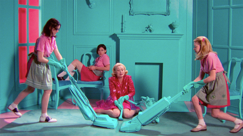 Three girls in pink tops and green skirts vacuum around a woman in a shiny pink coat, all in a monochrome turquoise room with matching furniture and props.