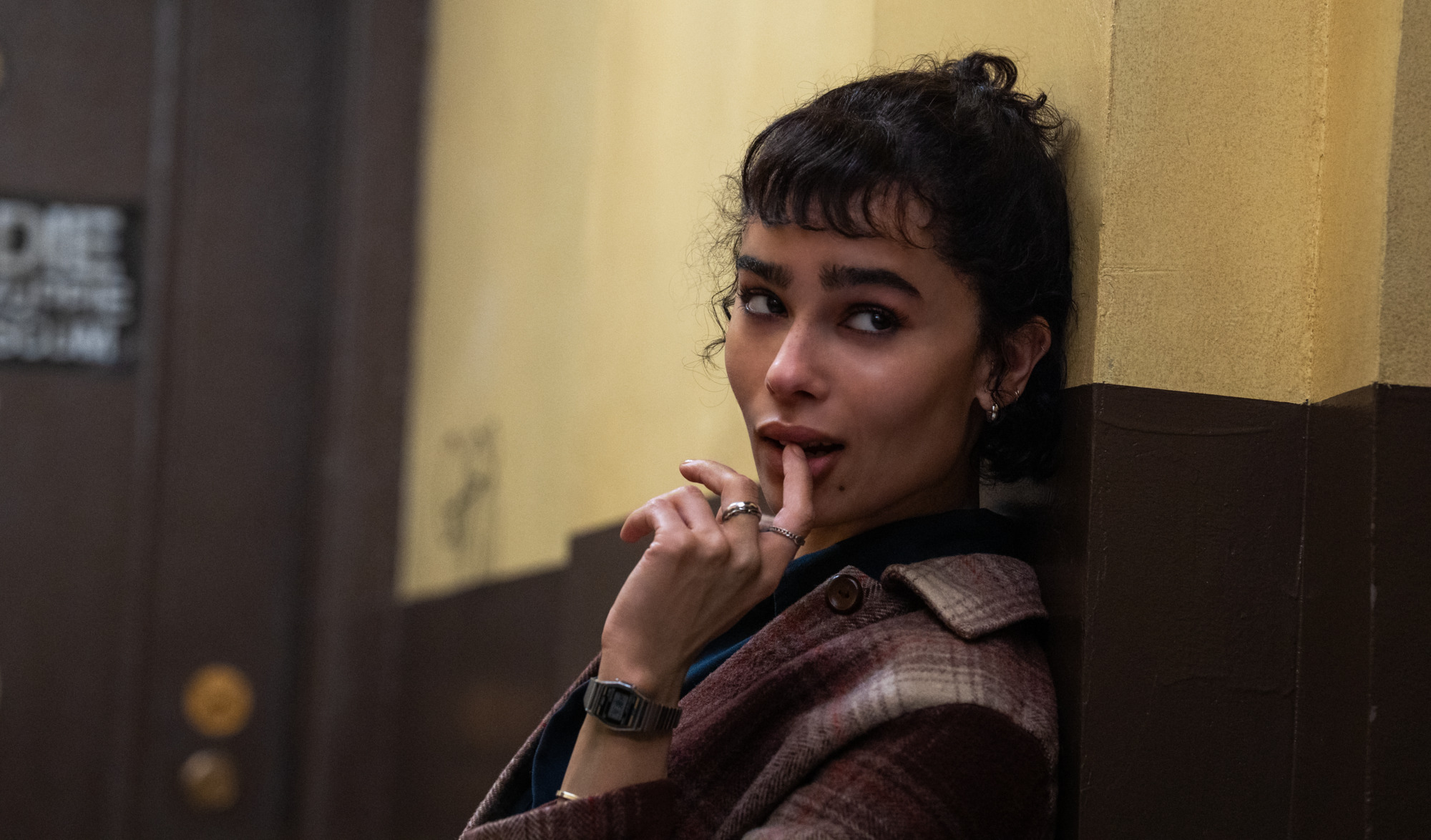 A woman with short curly hair leans against a yellow and brown wall, looking to the side with her finger resting on her lips.