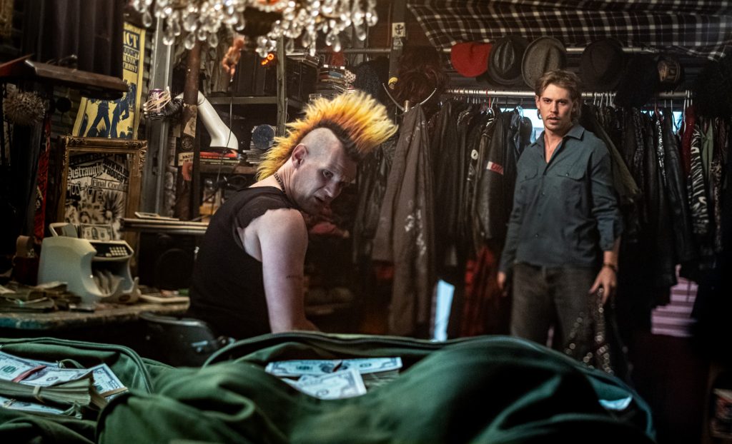A man with a yellow mohawk sits at a cluttered table with cash, while another man stands in a dimly lit room filled with clothing and hats.