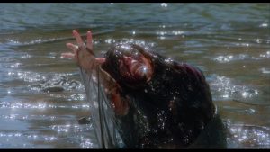A person covered in dark, oily sludge reaches upward while partially submerged in shallow water.