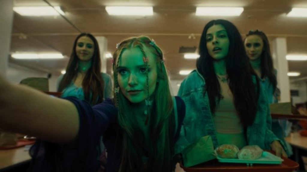 Four young women with food trays stand in a cafeteria. The woman in front reaches forward, illuminated by greenish light, while the others look toward the camera.