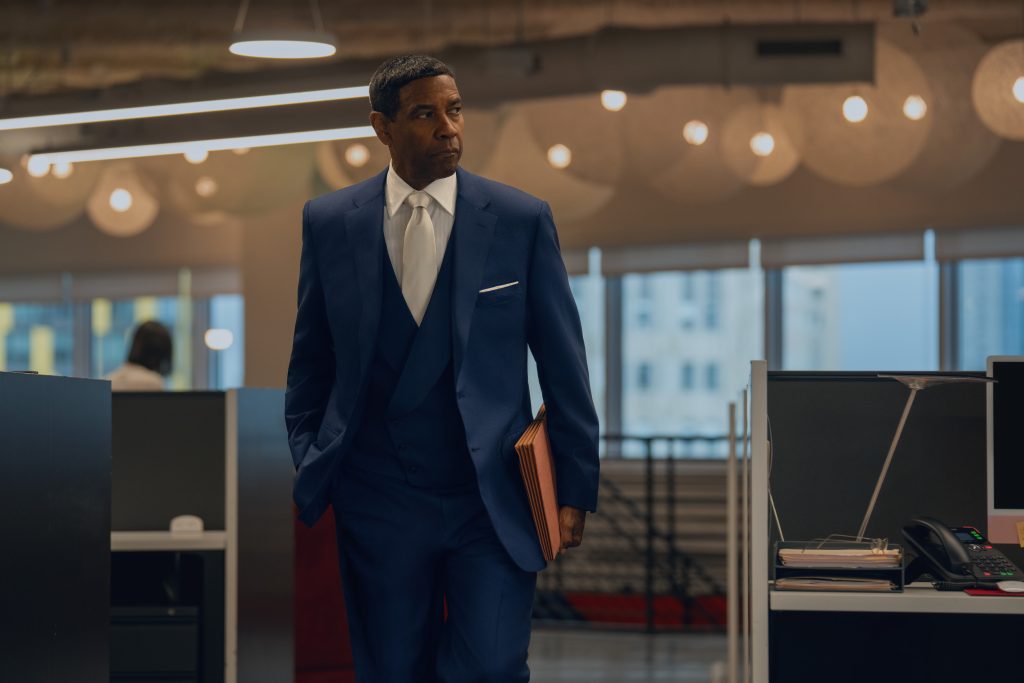 A man in a blue suit holding a folder walks through a modern office space with desks and overhead lights.