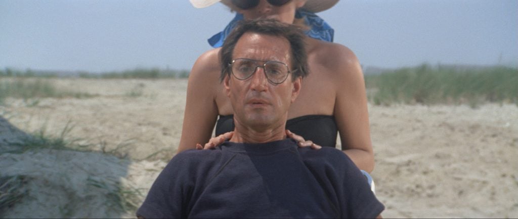 A man wearing glasses sits on a beach with a concerned expression while a woman stands behind him, resting her hands on his shoulders.