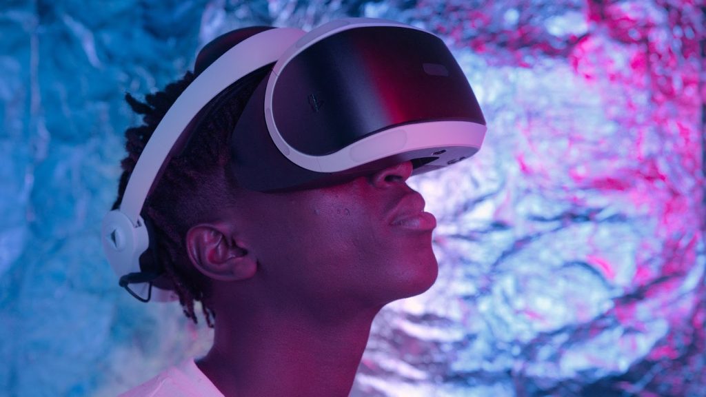 A person wearing a virtual reality headset looks upward, illuminated by pink and blue lights against a metallic, textured background.