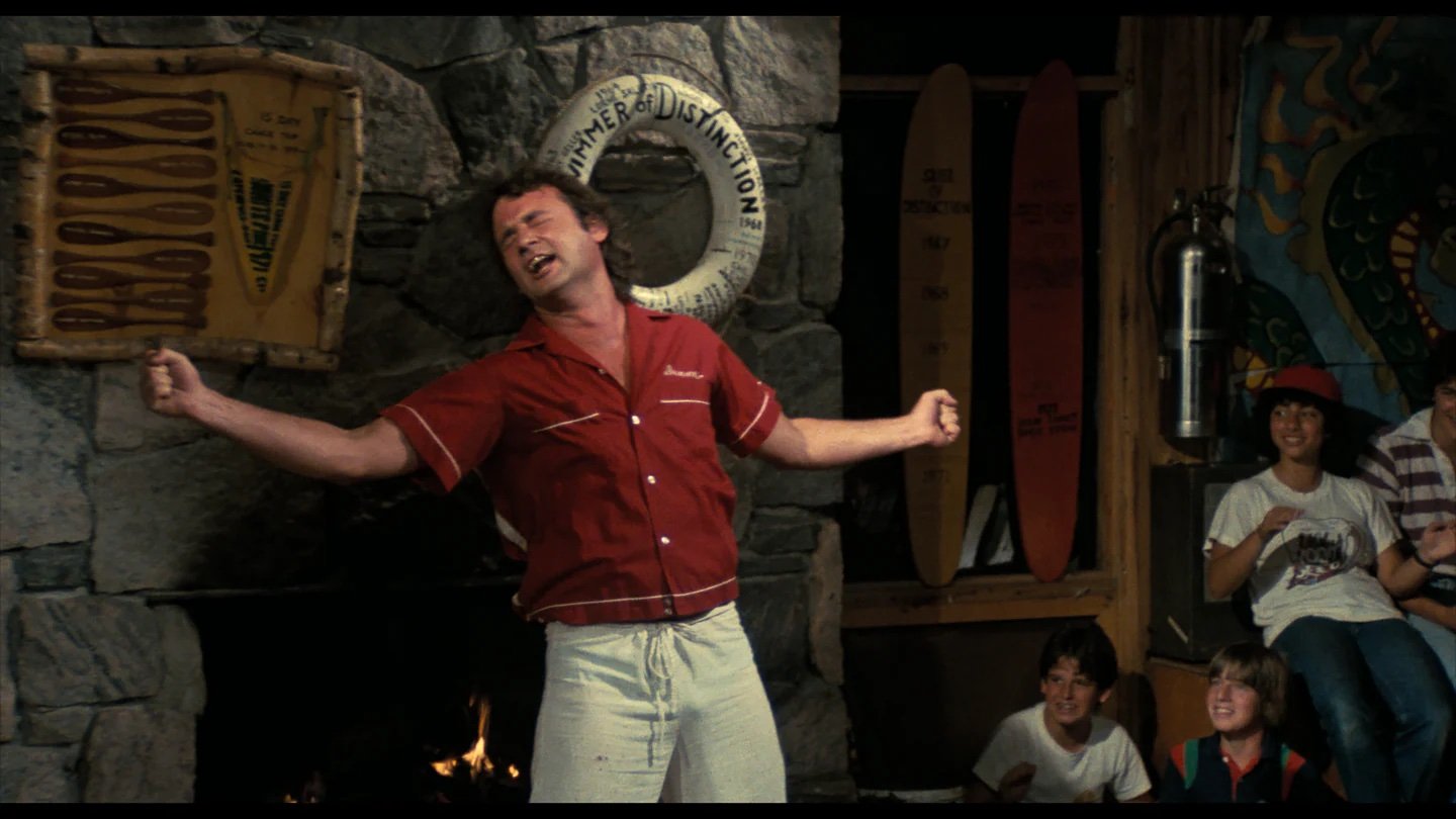 A man in a red shirt and white shorts stands with arms outstretched, performing enthusiastically in front of a stone wall as several people watch and smile.