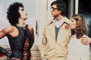 Three people stand together; the person on the left wears a black sequined outfit, while the two on the right wear light-colored clothing and look toward the left person.