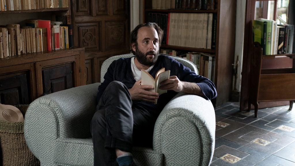 A man with a beard sits in an armchair reading a book, surrounded by bookshelves in a cozy, well-lit room.