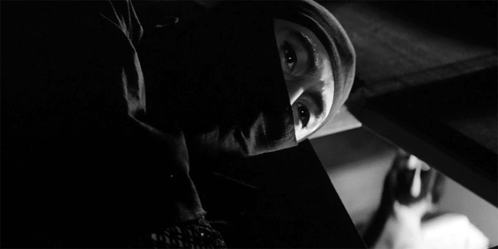 A person wearing a mask peers up from behind a dark surface, their face partially illuminated by light from an open drawer or compartment.