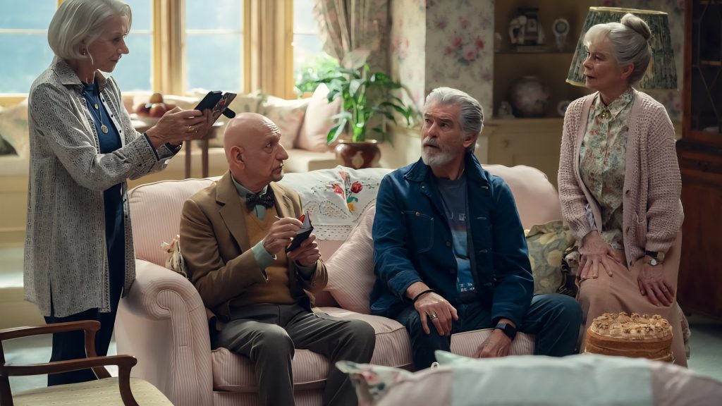 Four elderly people sit and stand in a cozy, well-lit living room with floral decor. Two women stand, one holding a phone, while two men sit on a sofa, talking. A pie rests on the table.