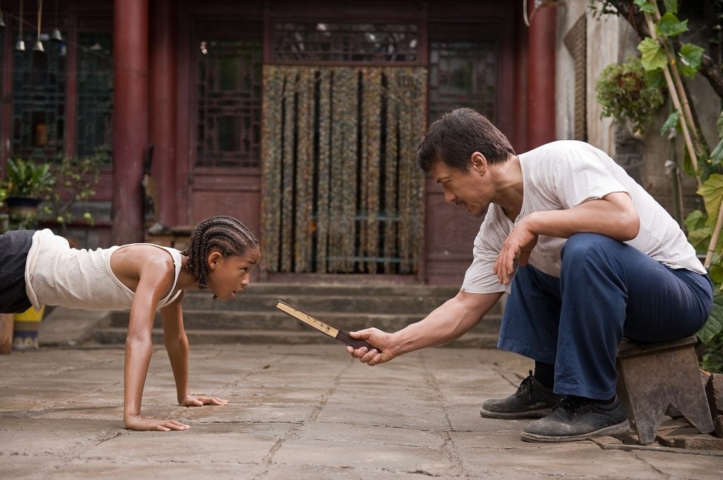 A young girl does push-ups while an older man sits in front of her, holding a wooden board near her face in an outdoor courtyard.