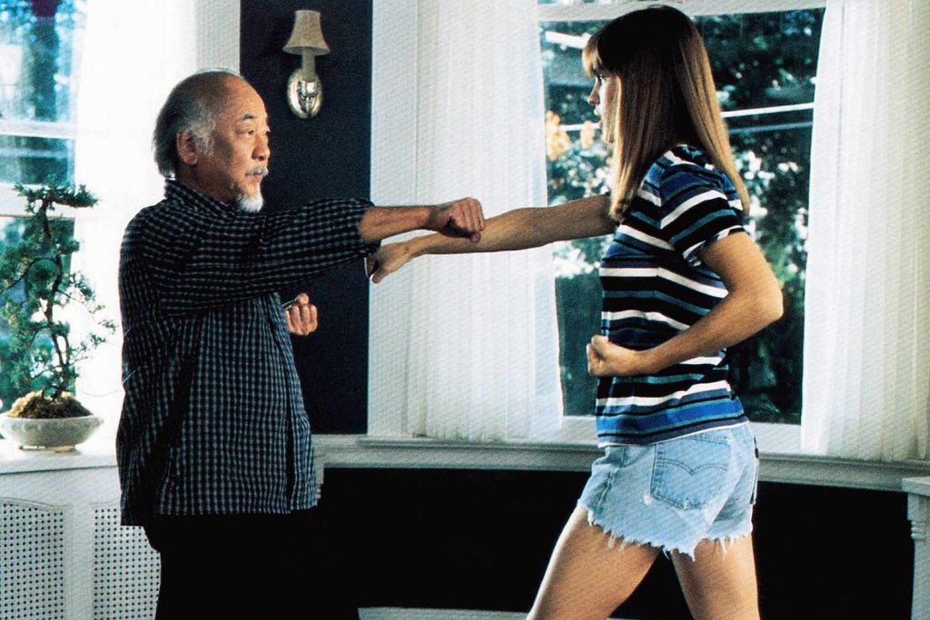 An older man instructs a young woman in a striped shirt and denim shorts on a punching technique inside a sunlit room.